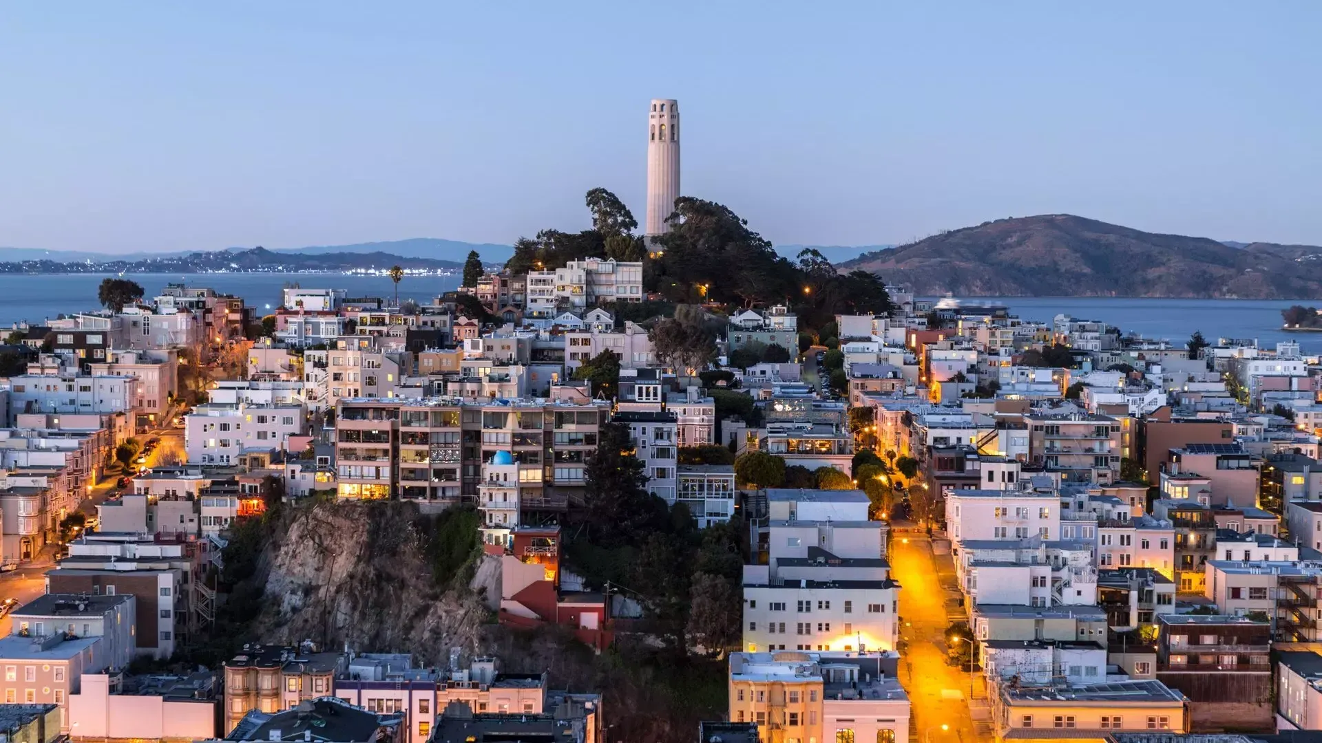 La Torre Coit de San Francisco al anochecer, 与灯火通明的街道在它前面和贝博体彩app湾在它后面.