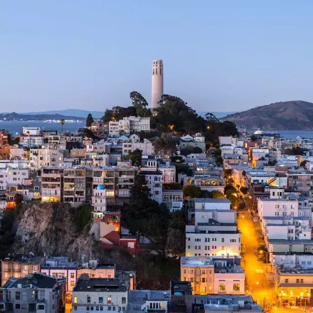 Coit Tower, em São Francisco, ao anoitecer, com ruas iluminadas à sua frente e a Baía de São Francisco atrás.